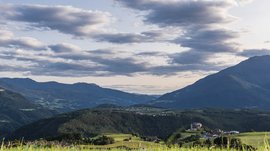 Rodeneck – for a wonderful walking holiday in Italy The picture shows a wide landscape with gentle, wooded hills, over which lies a slightly cloudy sky. In the foreground, green meadows are visible, while in the distance a village nestles into the hills.