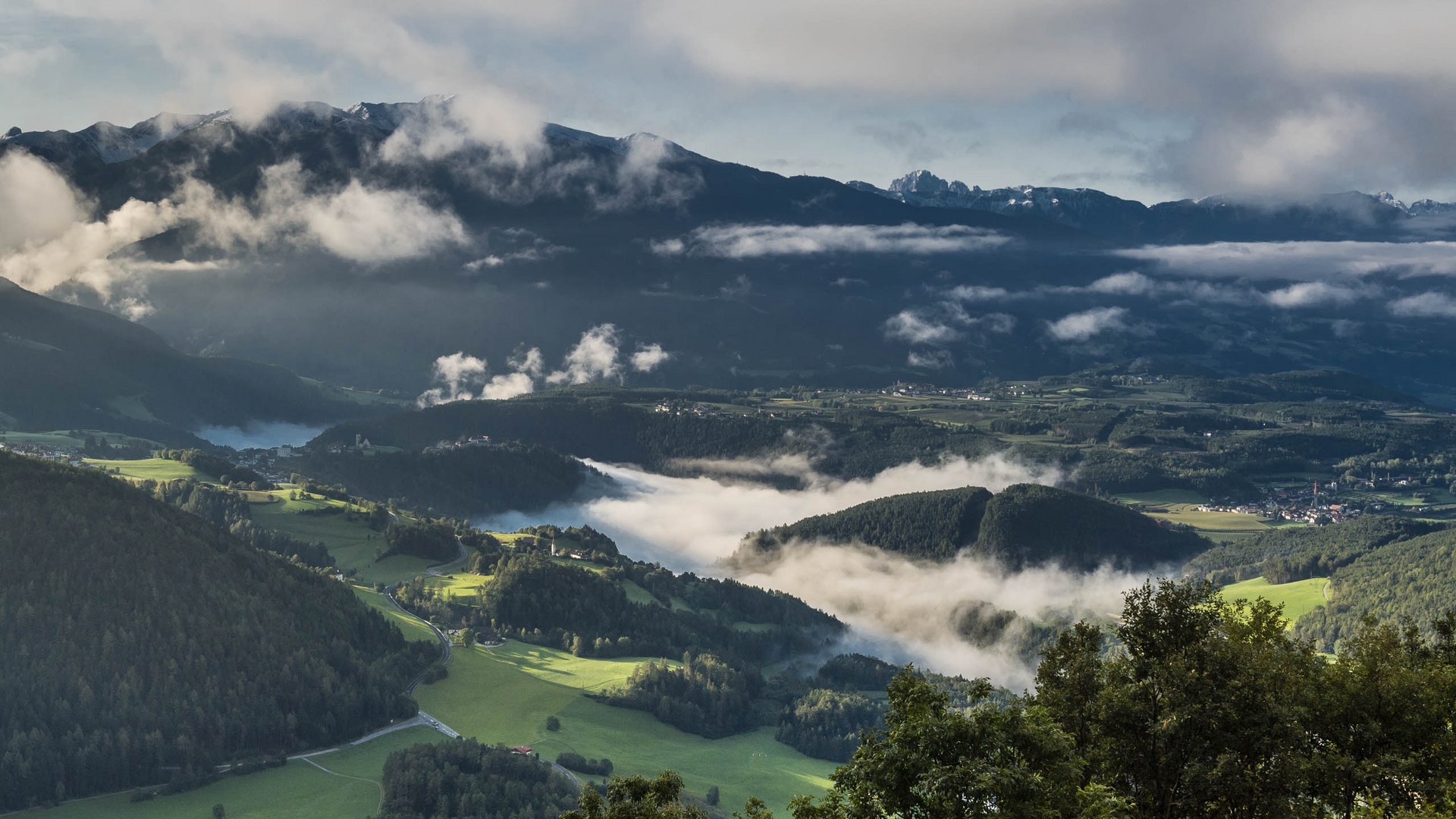 Rodeneck – for a wonderful walking holiday in Italy The picture shows a vast, green landscape with gentle hills and valleys, through which misty clouds drift. In the background, dark mountains rise into the sky, partially covered by clouds, while the low sun illuminates the scene.