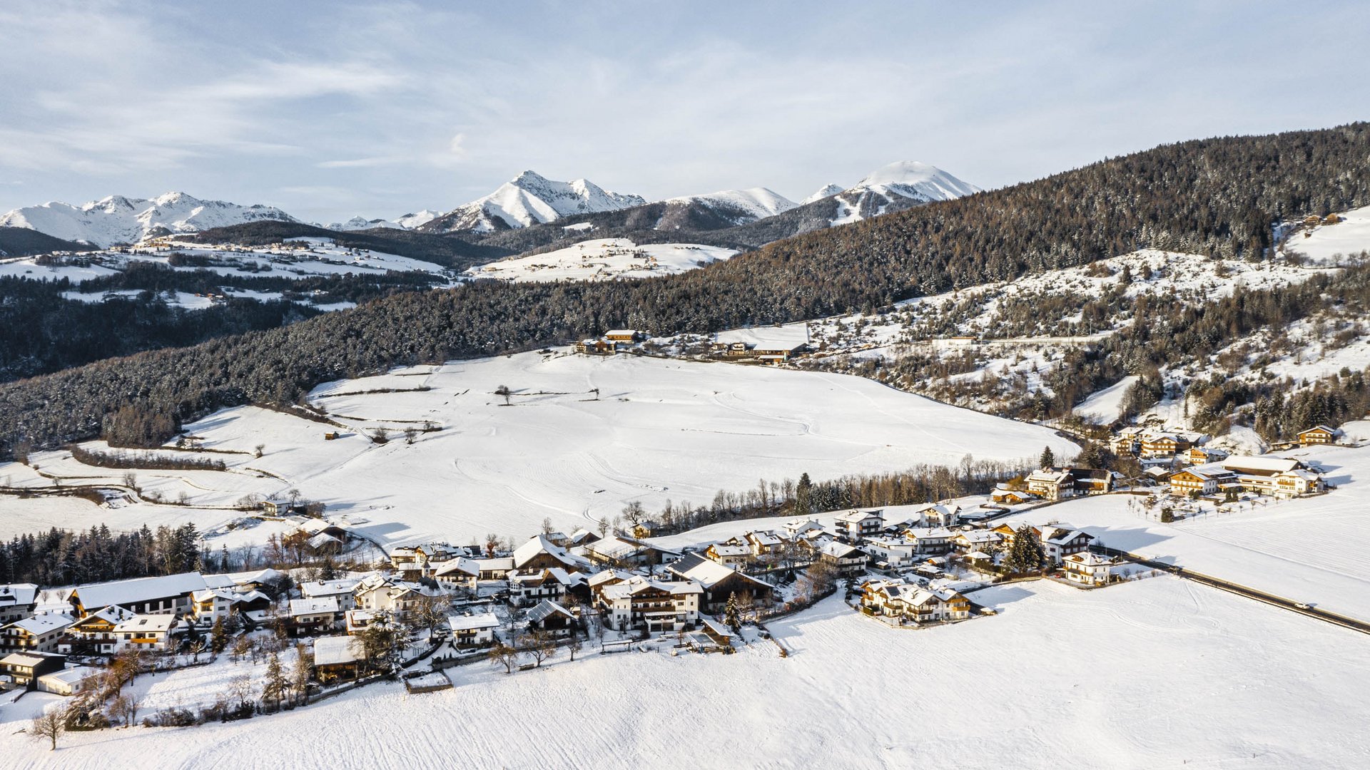 Rodeneck – for a wonderful walking holiday in Italy The picture shows a snowy landscape with a village stretching out in the foreground. In the background, forested hills and snow-covered mountains can be seen beneath a clear sky. The houses of the village are partially covered with snow, and the scene feels calm and peaceful.