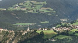 Rodeneck – for a wonderful walking holiday in Italy The picture shows an expansive landscape with a castle and a church perched on a rock. In the background, green fields, forests, and small villages stretch out in a hilly environment.