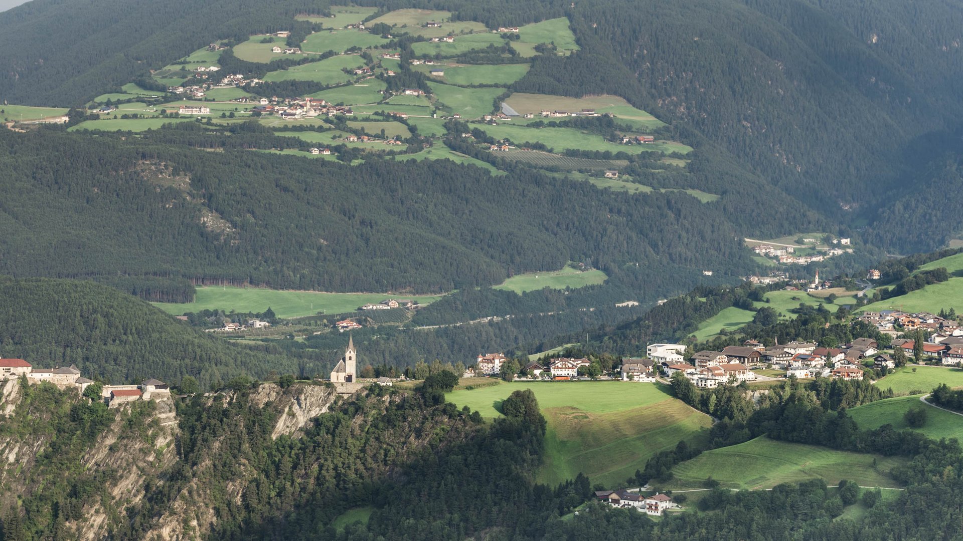 Rodeneck – for a wonderful walking holiday in Italy The picture shows an expansive landscape with a castle and a church perched on a rock. In the background, green fields, forests, and small villages stretch out in a hilly environment.