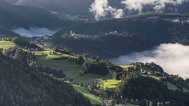 Rodeneck – for a wonderful walking holiday in Italy The picture shows a hilly landscape with green meadows and forests, through which a road winds. In the background, a church and a small village can be seen, surrounded by fog that moves through the valleys.