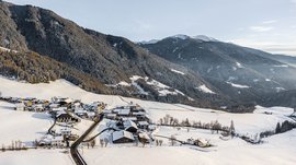 Rodeneck – for a wonderful walking holiday in Italy The picture shows a small snowy village located in a hilly landscape. In the background, wooded mountains rise, also covered by a thin layer of snow. The scene appears calm and wintry, with a clear sky and scattered buildings lying in the snow.