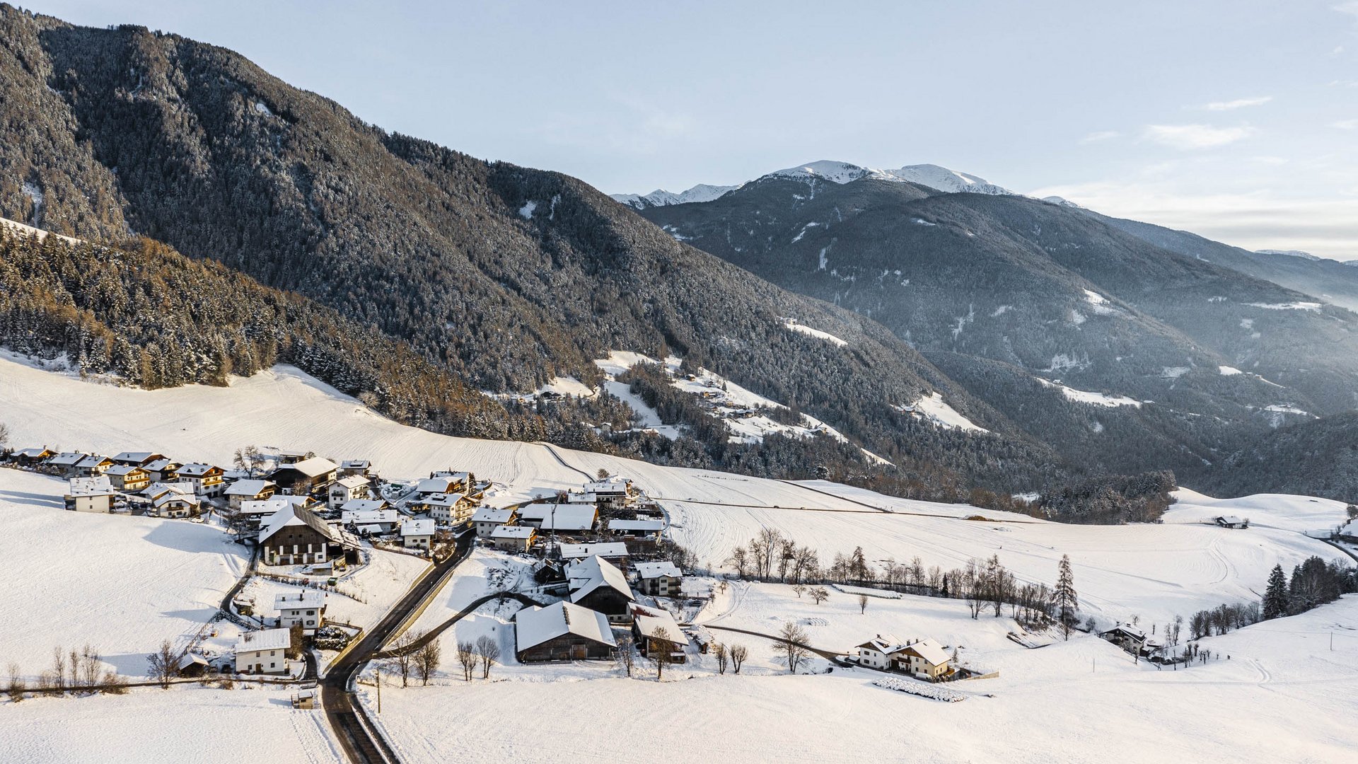 Rodeneck – for a wonderful walking holiday in Italy The picture shows a small snowy village located in a hilly landscape. In the background, wooded mountains rise, also covered by a thin layer of snow. The scene appears calm and wintry, with a clear sky and scattered buildings lying in the snow.