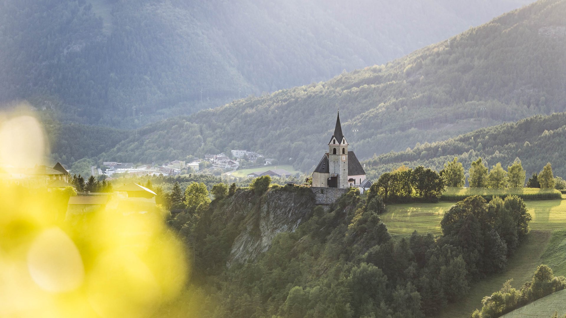 Rodeneck – for a wonderful walking holiday in Italy The picture shows a church on a rock, surrounded by green trees and meadows, with mountains in the background. The sun casts soft light on the scene, making the landscape peaceful and idyllic.