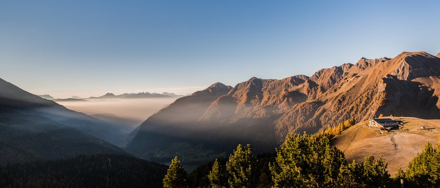 The Gitschberg webcams The picture shows a picturesque mountain landscape at sunrise or sunset, with deep valleys covered in fog and sunlit mountain slopes glowing in warm orange tones. On a ridge to the right in the image stands a mountain hut, surrounded by grassy hills and coniferous trees.