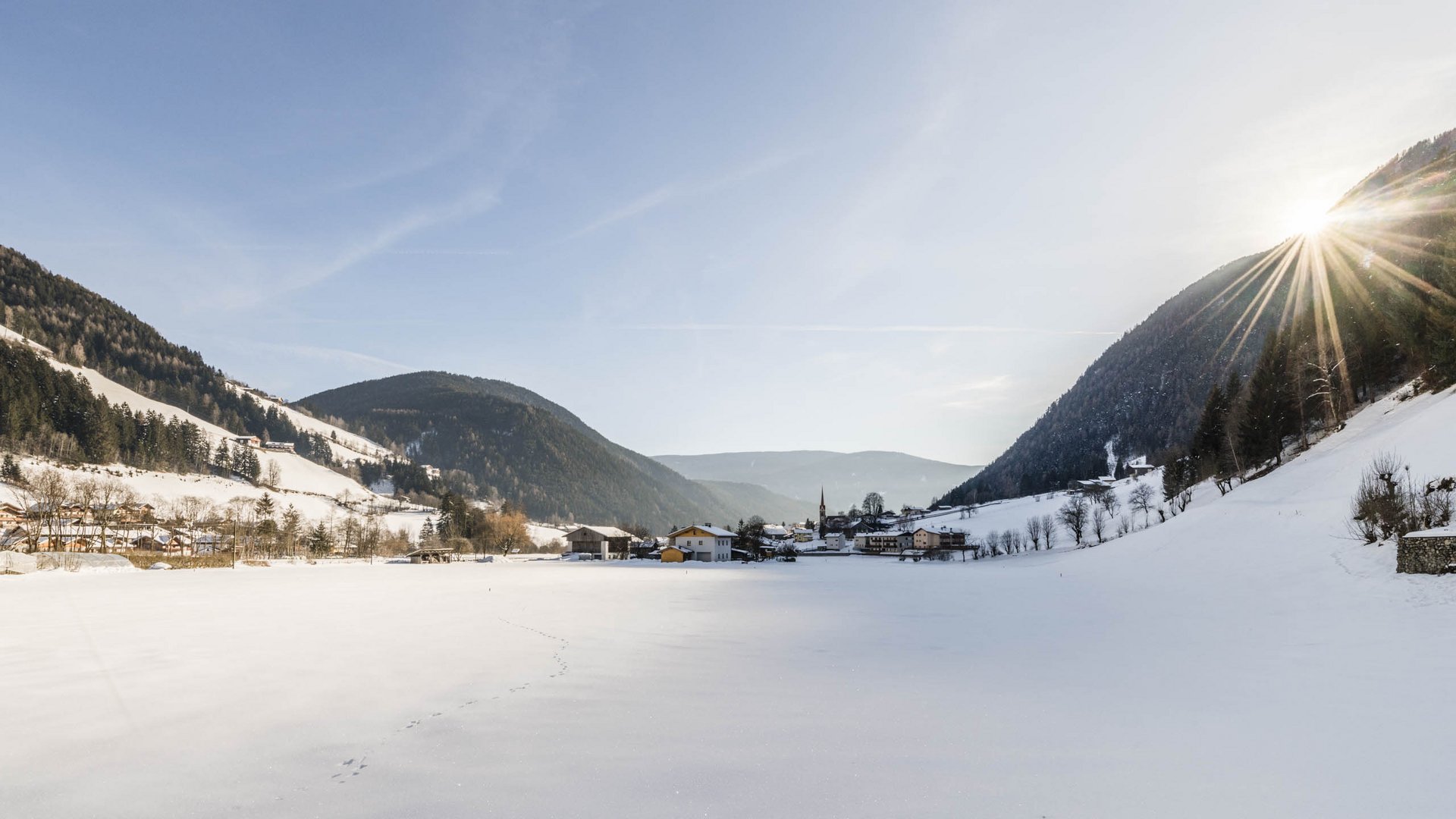 Pfunders: urig und idyllisch Das Bild zeigt eine verschneite Winterlandschaft in einem Tal. In der Ferne ist ein kleines Dorf zu sehen, das von schneebedeckten Hügeln und Wäldern umgeben ist. Die Sonne scheint hell und strahlt über den Bergkamm rechts im Bild.