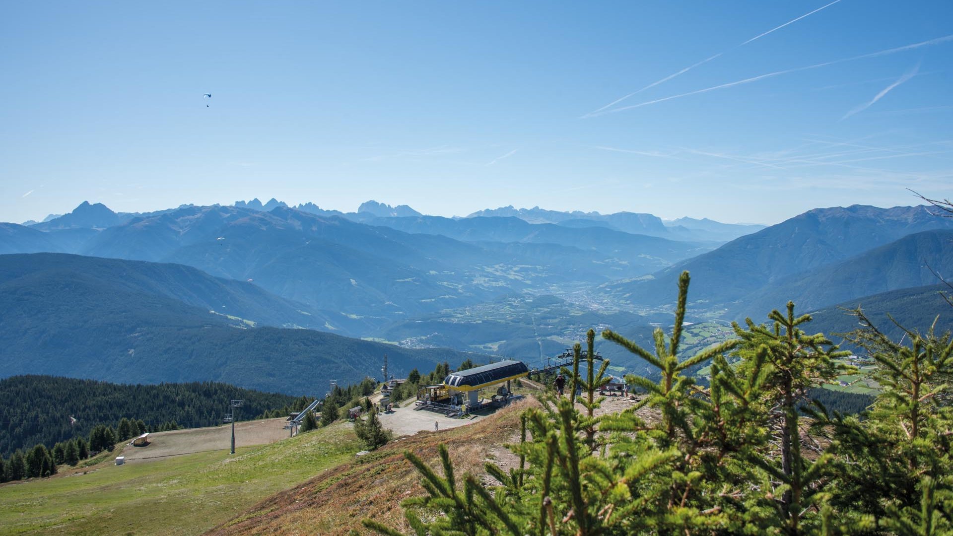 Gitschberg: Najít hotely Obrázek ukazuje rozsáhlou horskou krajinu s jasně modrou oblohou. V popředí jsou některé jedle a níže je vidět horská stanice s lanovkou. V pozadí se až k horizontu táhnou hory a údolí.