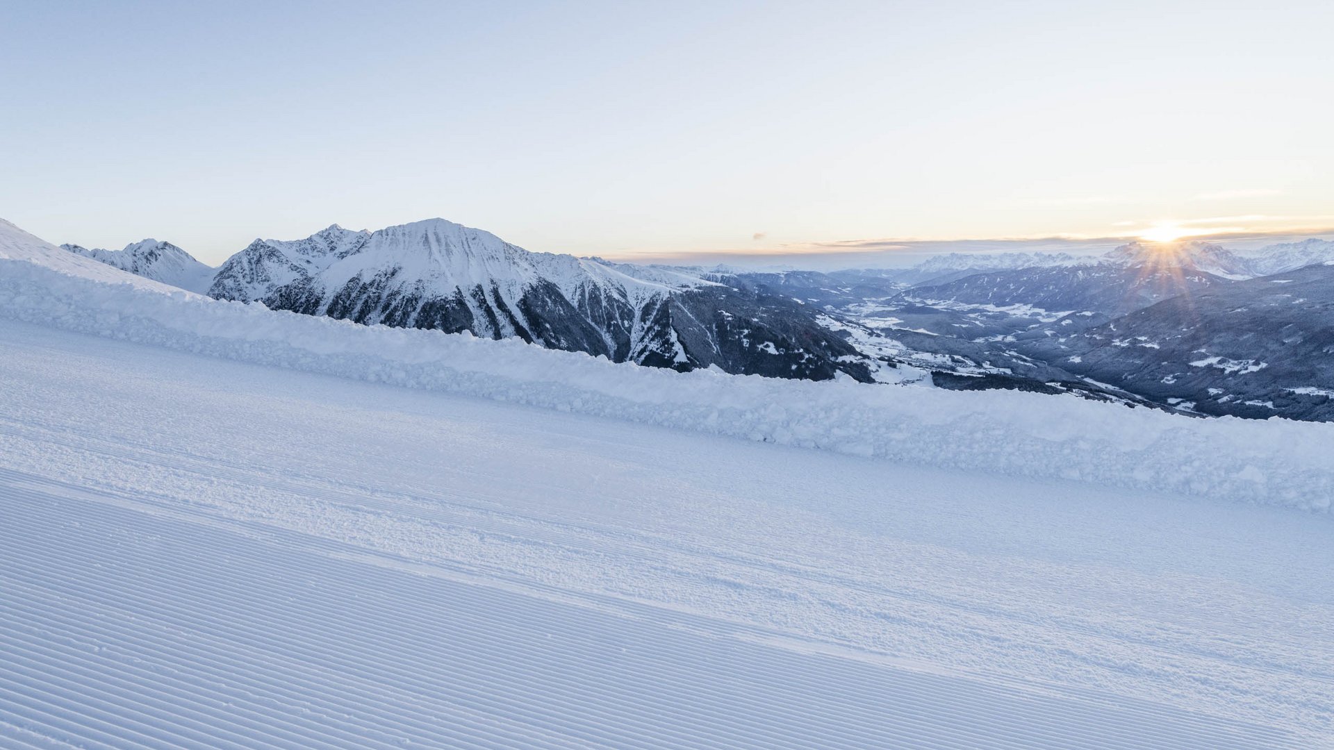 Firn, Ski & Schnee Frisch präparierte Skipiste mit verschneiten Bergen im Sonnenaufgang