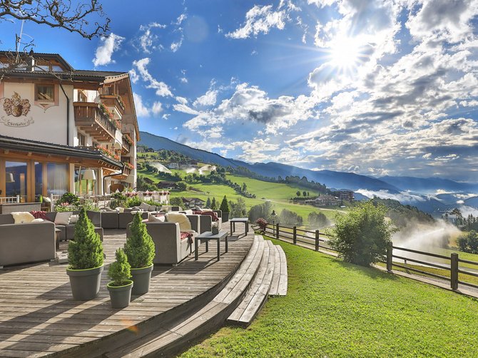 Willkommen in der Ferienregion Gitschberg Jochtal! Terrasse eines Gasthauses mit grüner Berglandschaft und Sonne am Himmel