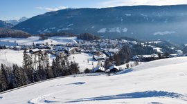 Langlaufen in Terenten De foto toont een besneeuwd berglandschap met een klein dorp in de vallei, omgeven door heuvels en bergen. De lucht is helder en blauw, en op de voorgrond zijn sporen in de sneeuw op een zachte helling te zien.