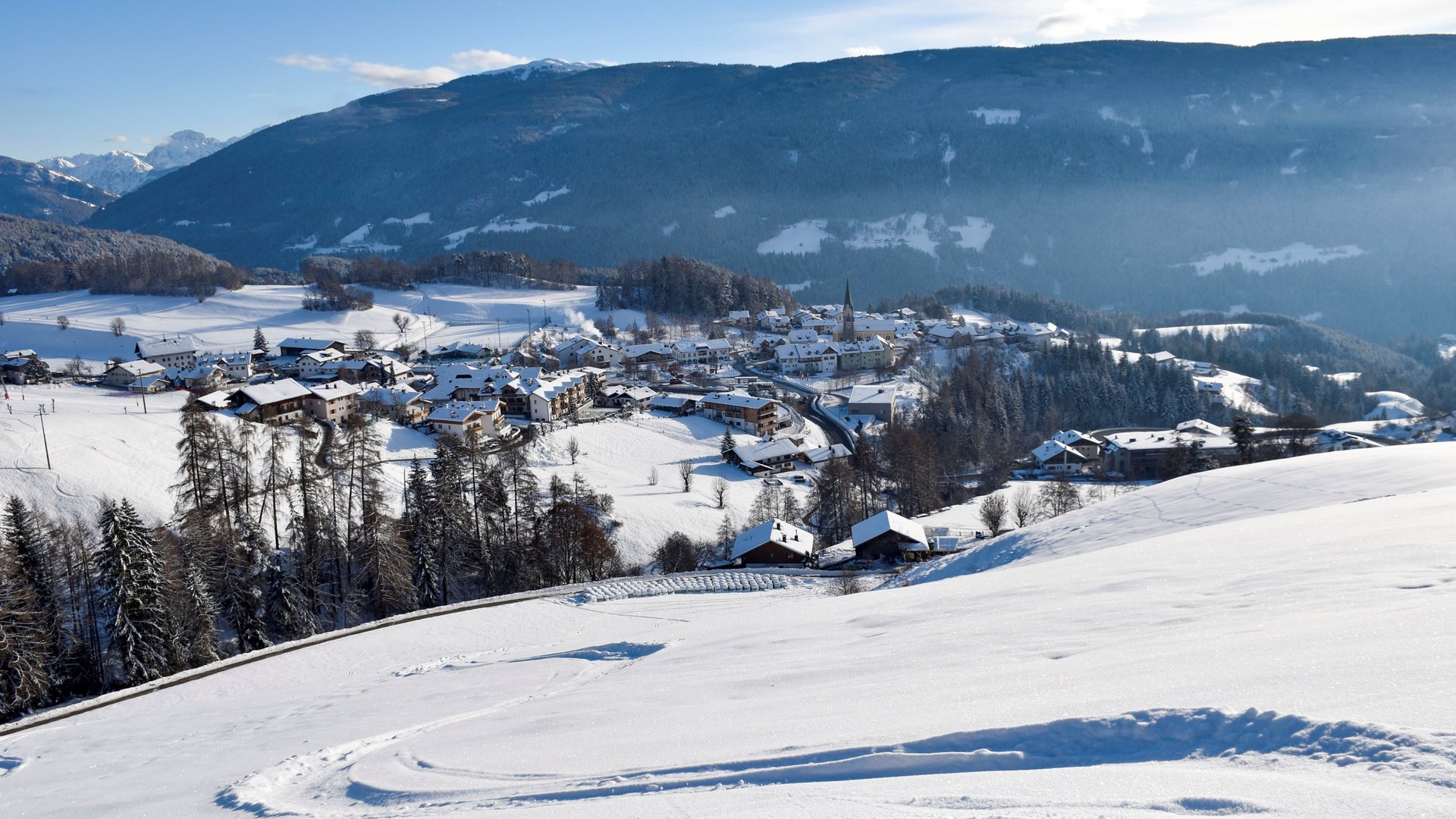 Langlaufen in Terenten De foto toont een besneeuwd berglandschap met een klein dorp in de vallei, omgeven door heuvels en bergen. De lucht is helder en blauw, en op de voorgrond zijn sporen in de sneeuw op een zachte helling te zien.