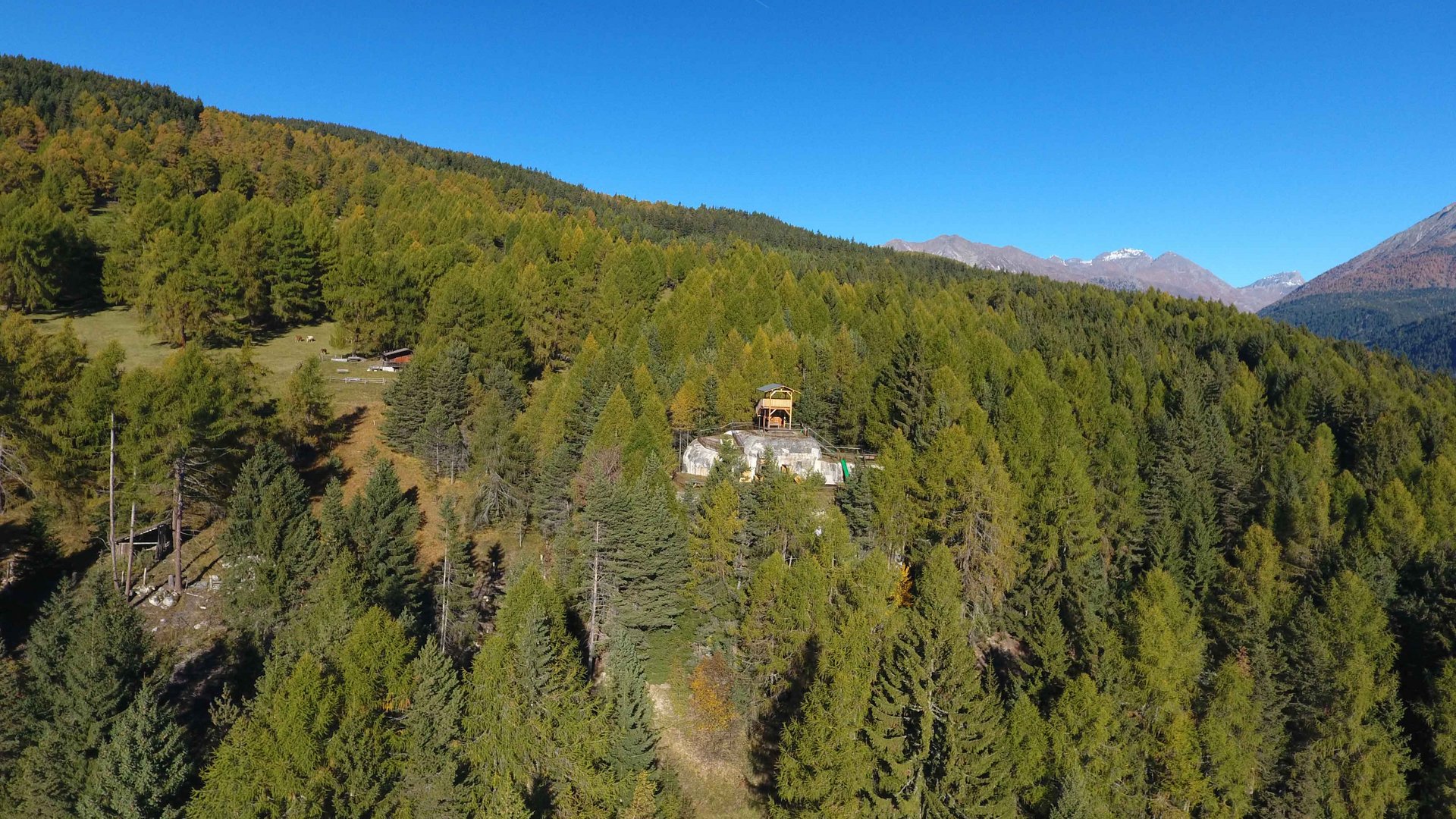 The recreation area in Spinges, South Tyrol The picture shows a small building located on a wooded hill, surrounded by dense forest and mountains in the background. The sky is clear and blue, and the terrain is covered with many green trees.