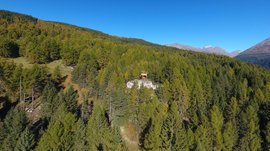 The recreation area in Spinges, South Tyrol The picture shows a small building located on a wooded hill, surrounded by dense forest and mountains in the background. The sky is clear and blue, and the terrain is covered with many green trees.