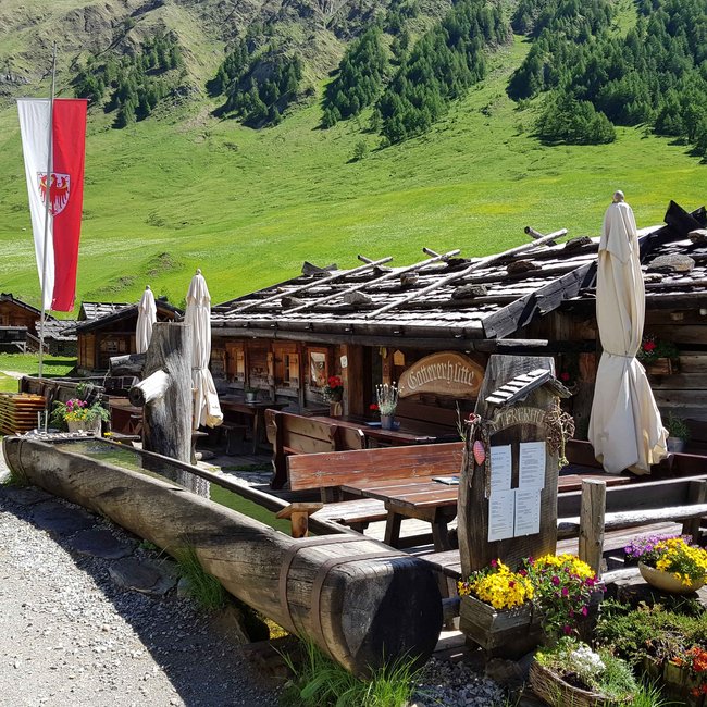 Gattererhütte Obrázek ukazuje rustikální dřevěnou chatku s venkovním prostorem vybaveným dřevěnými stoly, lavicemi a srolovanými slunečníky, obklopenou bujnou zelenou alpskou krajinou a červeno-bílou vlajkovou tyčí.