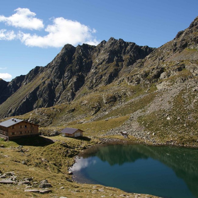 Tiefrastenhütte The picture shows a mountain hut next to a calm, clear mountain lake in a rocky alpine landscape. Steep, rugged mountains rise in the background under a blue sky with scattered clouds.