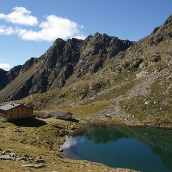 Südtiroler Almkost Das Bild zeigt eine Berghütte neben einem ruhigen, klaren Bergsee in einer felsigen, alpinen Landschaft. Steile, zerklüftete Berge erheben sich im Hintergrund unter einem blauen Himmel mit vereinzelten Wolken.