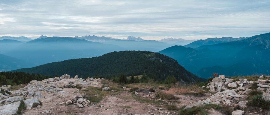 The Gitschberg webcams The picture shows a mountain landscape with several layers of hills and mountains stretching into the distance. The foreground consists of a rocky terrain, while in the background wooded hills and more distant mountain ranges can be seen under a cloudy sky.