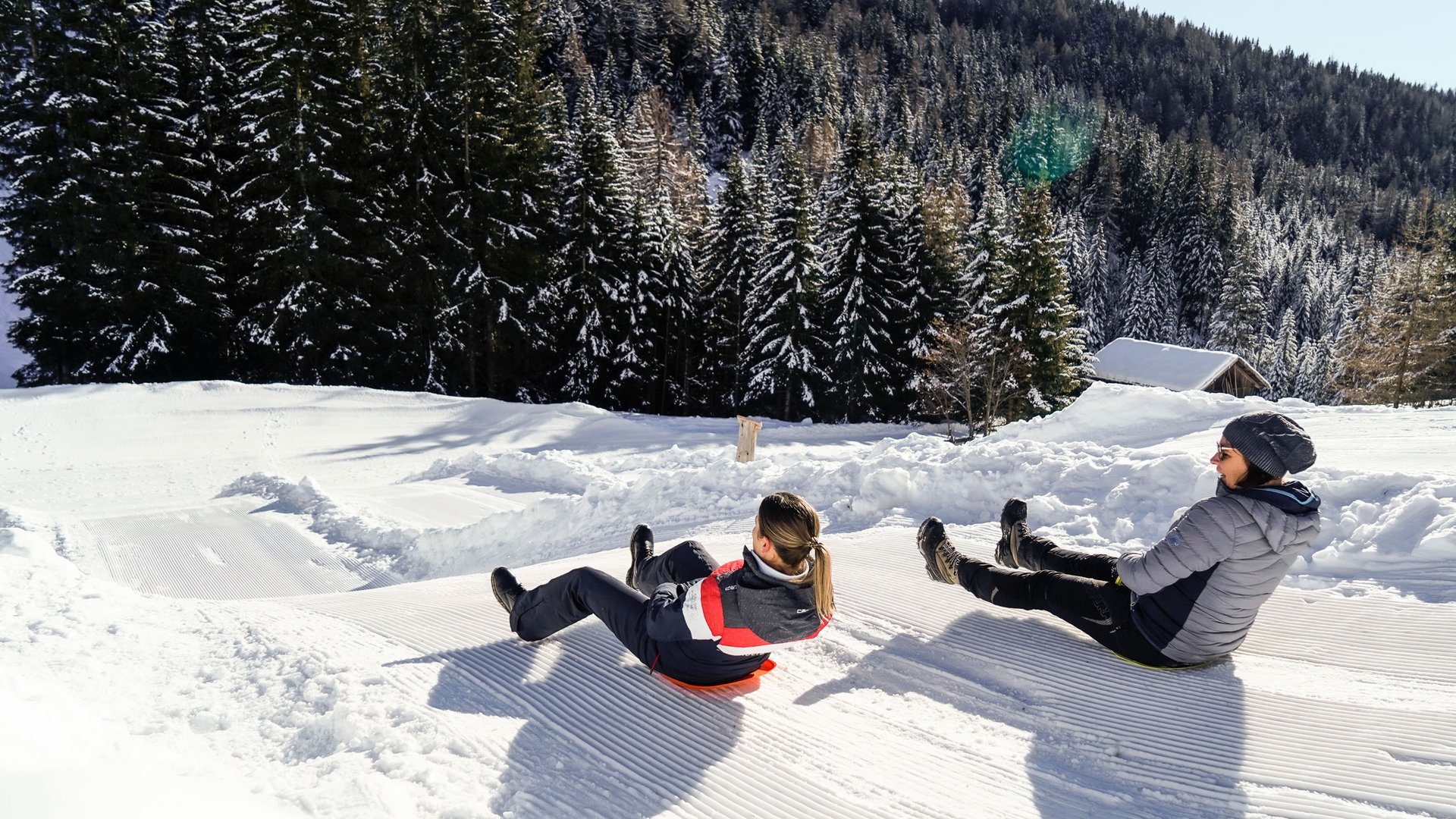 The Altfasstal valley, Meransen The picture shows two people sledding down a groomed snow slope. They are wearing winter clothing and are surrounded by snow-covered trees and a wooded hill in the background. It is a clear, sunny day.