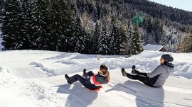 The Altfasstal valley, Meransen The picture shows two people sledding down a groomed snow slope. They are wearing winter clothing and are surrounded by snow-covered trees and a wooded hill in the background. It is a clear, sunny day.