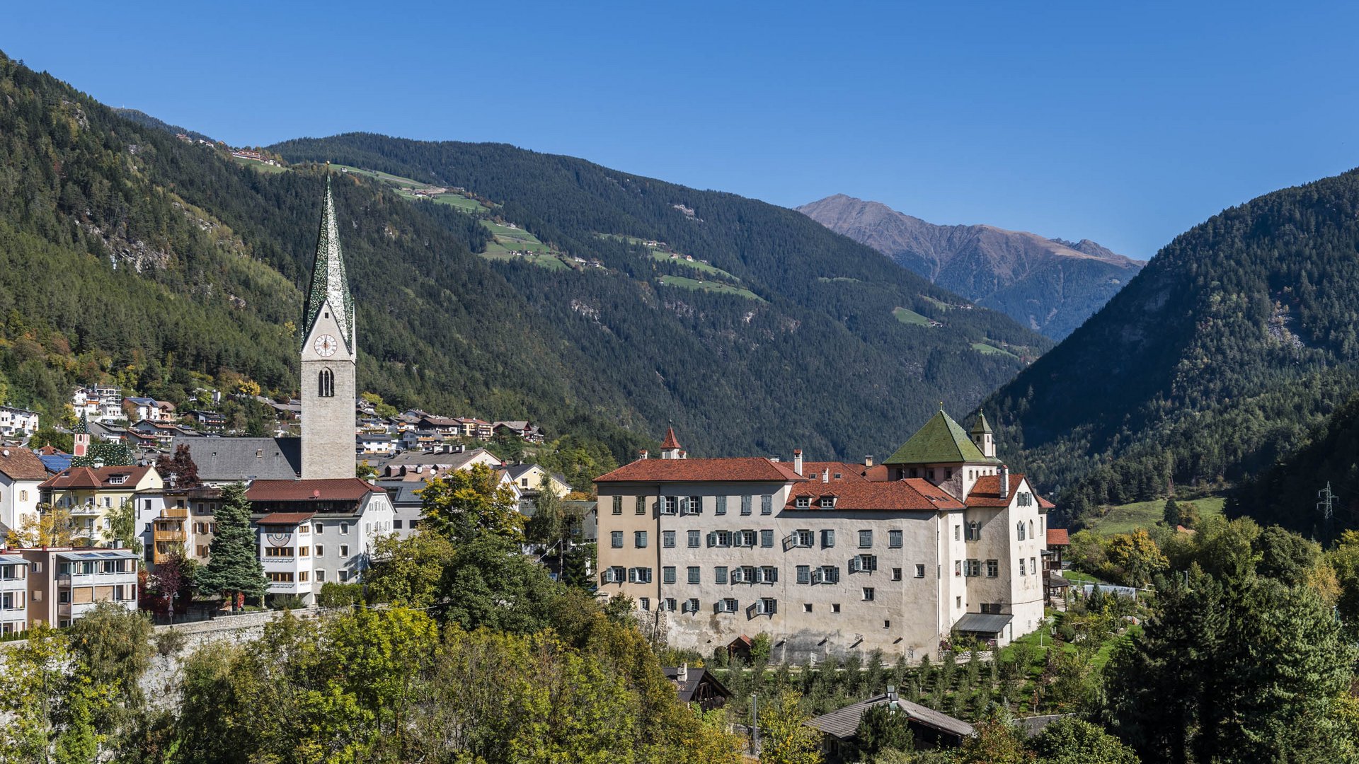 Mühlbach im Pustertal Das Bild zeigt eine malerische Kleinstadt in einem Tal, umgeben von bewaldeten Hügeln und Bergen. In der Mitte steht eine Kirche mit einem hohen, spitzen Turm, und daneben befindet sich ein großes Gebäude mit roten Dächern und vielen Fenstern.