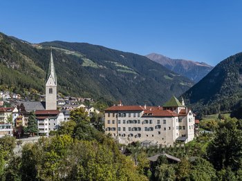 Mühlbach in the Pustertal valley The picture shows a picturesque small town in a valley, surrounded by wooded hills and mountains. In the center is a church with a tall, pointed tower, and next to it is a large building with red roofs and many windows.