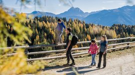Alpská pastvsina Rodenecker-Lüsner Alm Obrázek ukazuje rodinu na túře po stezce v horách. Dítě balancuje na dřevěném plotě, zatímco otec drží jeho ruku, a v pozadí jsou vidět barevné podzimní stromy a hory.