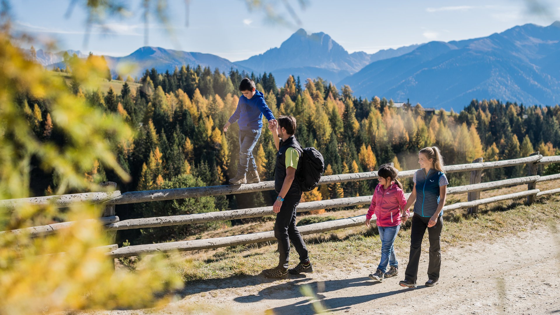 Alpská pastvsina Rodenecker-Lüsner Alm Obrázek ukazuje rodinu na túře po stezce v horách. Dítě balancuje na dřevěném plotě, zatímco otec drží jeho ruku, a v pozadí jsou vidět barevné podzimní stromy a hory.
