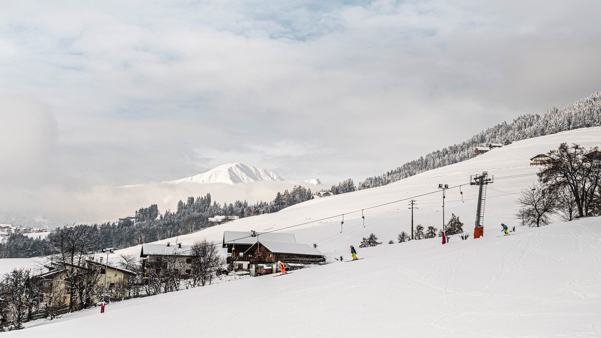 Skivakantie met kinderen in Zuid-Tirol/Terenten De afbeelding toont een met sneeuw bedekt landschap met een skilift en meerdere skiërs op een piste. Op de achtergrond zijn besneeuwde heuvels en bergen te zien, die gedeeltelijk door wolken worden bedekt.