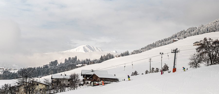The Gitschberg webcams The picture shows a snow-covered landscape with a ski lift and several skiers on a slope. In the background, snowy hills and mountains can be seen, partially obscured by clouds.