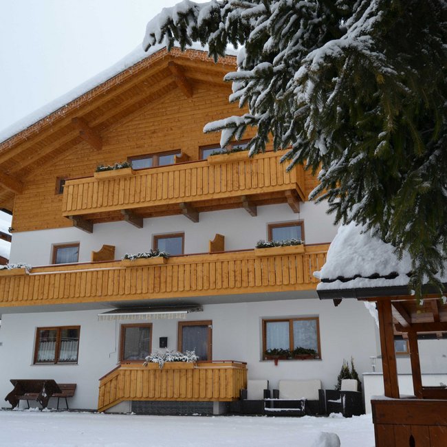 Selský šenk Heisla Dvoupodlažní budova s bílou fasádou a dřevěnými balkony je pokrytá sněhem. Před domem stojí zasněžená lavička a v popředí je vidět zasněžený strom.