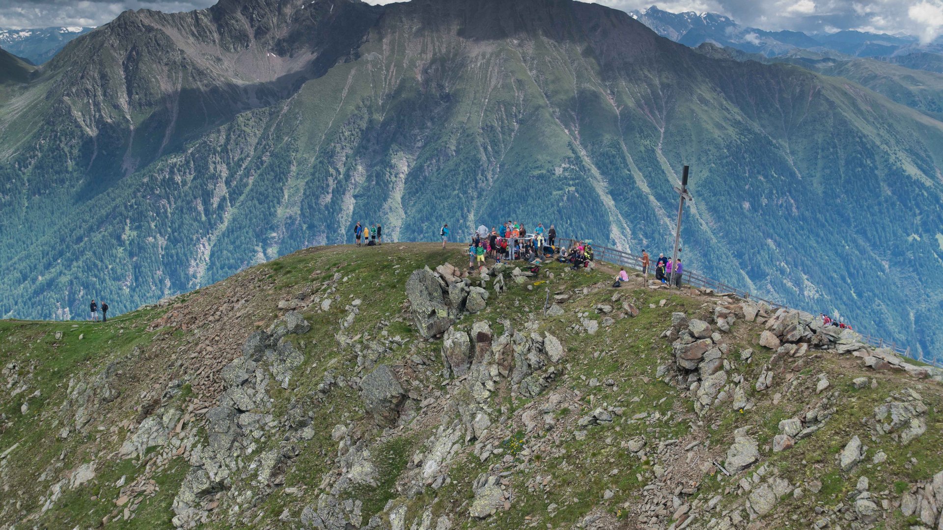 The Gitschberg panoramic platform in Meransen, Italy The picture shows a group of people standing on a rocky mountain peak. In the background, impressive green mountain ranges covered by clouds can be seen.