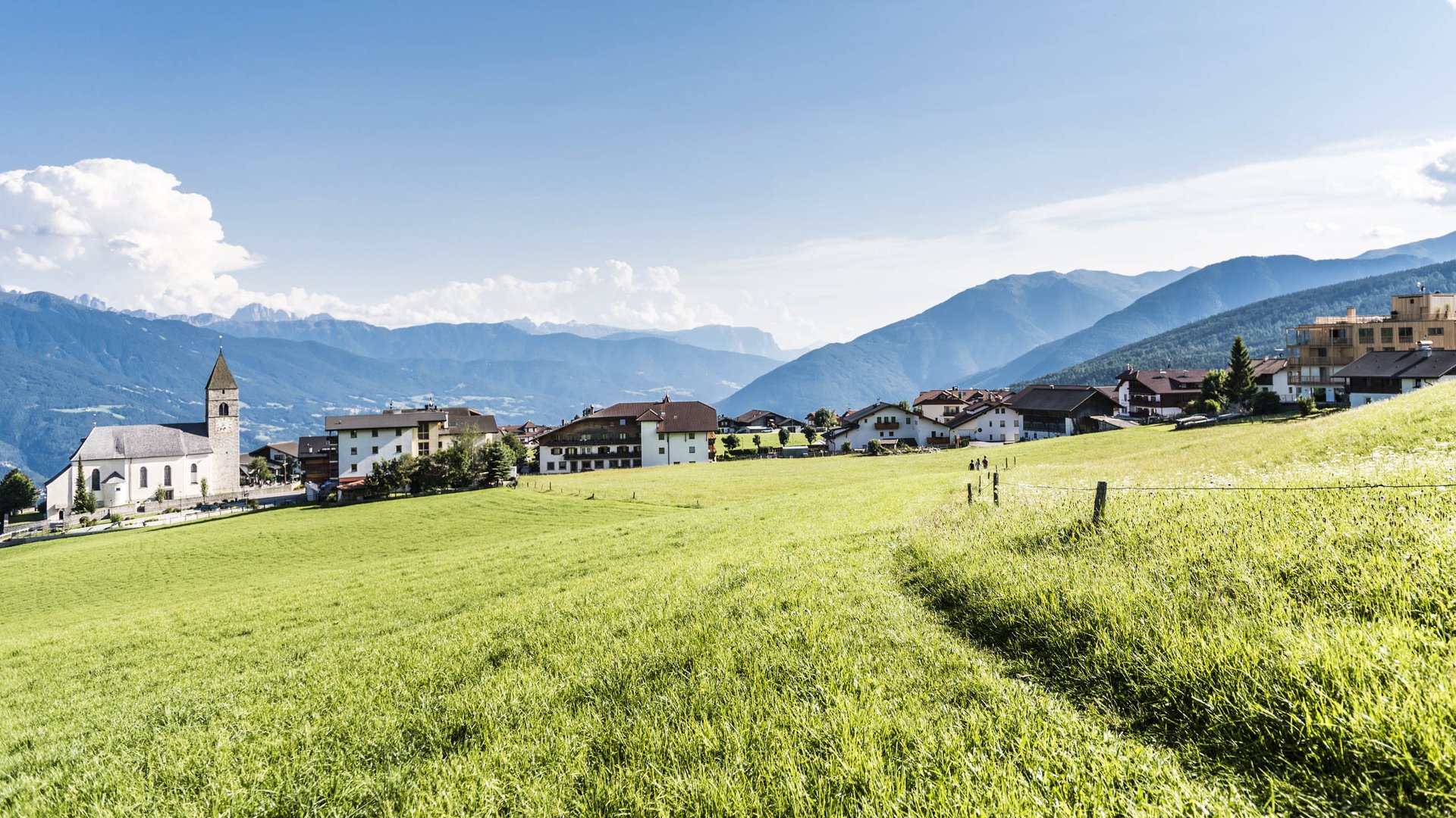 The mountain village of Meransen The picture shows a picturesque village with a church on the left side, surrounded by green meadows and scattered houses. In the background, forested mountains stretch out under a clear blue sky with few clouds.