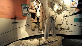 Loden Erlebniswelt in Vintl/Pustertal De afbeelding toont een tentoonstelling met skikleding uit de jaren 1930, gepresenteerd op etalagepoppen. Op de achtergrond zijn illustraties van skiërs en een oude poster te zien die de mode en de skisport uit die tijd weergeven. Een paar ski's ligt op de grond voor de kleding.
