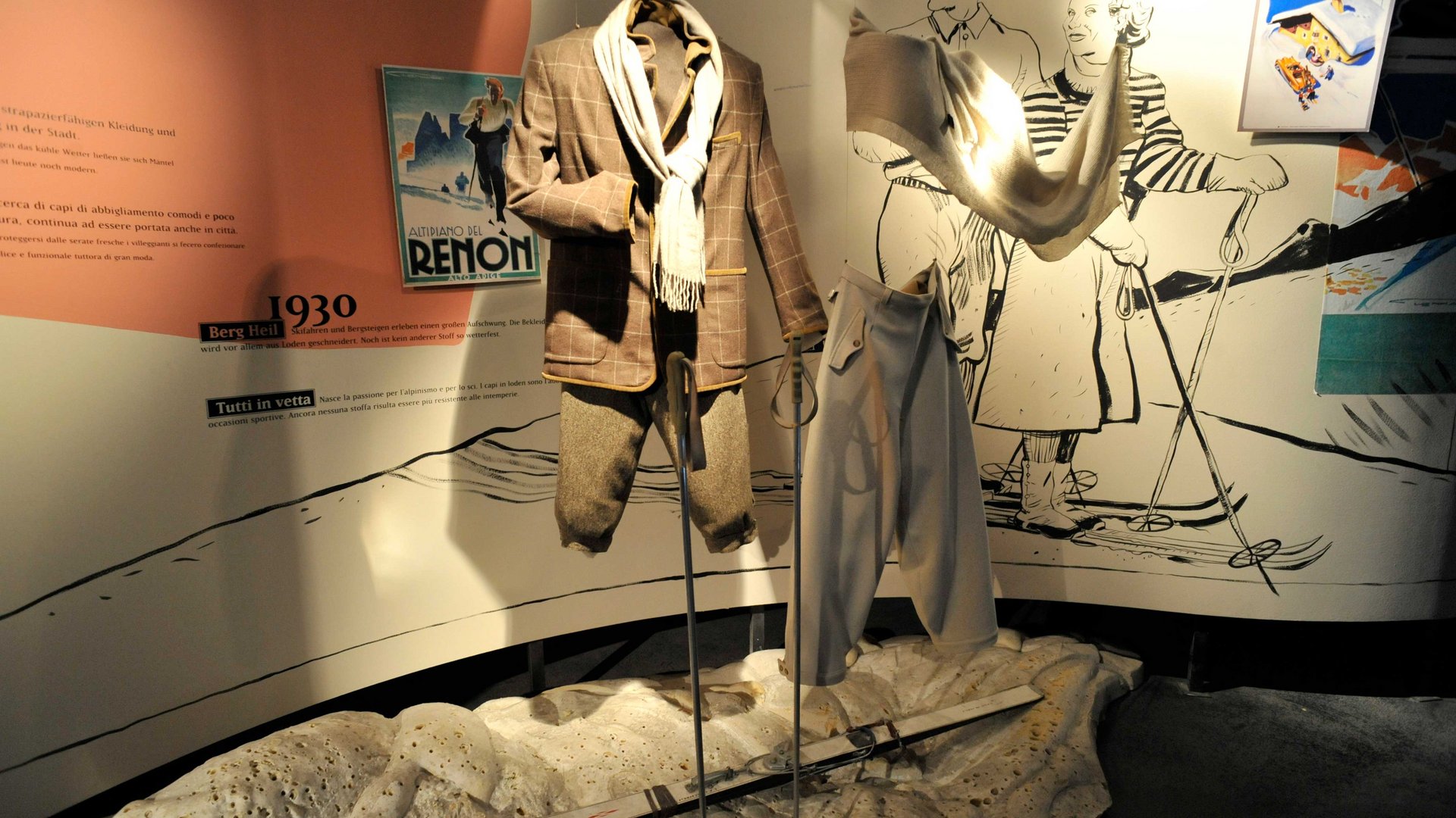 Loden Erlebniswelt in Vintl/Pustertal De afbeelding toont een tentoonstelling met skikleding uit de jaren 1930, gepresenteerd op etalagepoppen. Op de achtergrond zijn illustraties van skiërs en een oude poster te zien die de mode en de skisport uit die tijd weergeven. Een paar ski's ligt op de grond voor de kleding.