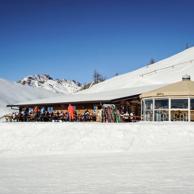 Nesselhütte De afbeelding toont een berghut in de sneeuw met een groot terras waarop meerdere mensen in skikleding zitten en ontspannen. Naast de hut staat een modern, glazen rond gebouw. De omgeving bestaat uit met sneeuw bedekte bergen onder een heldere, blauwe lucht.
