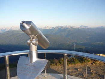 The mountain village of Meransen The picture shows a pair of fixed binoculars at a vantage point, pointed at an expansive mountain landscape. Mountain ranges and valleys can be seen in the background under a clear blue sky.