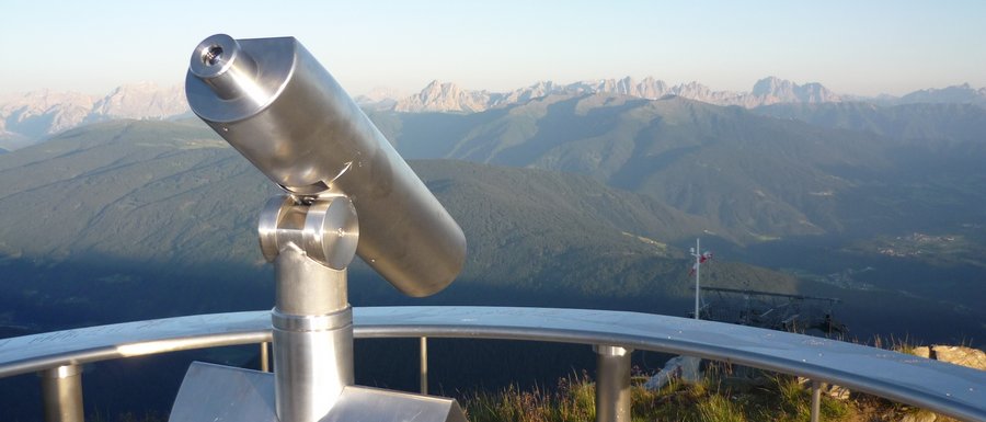 The Gitschberg webcams The image shows a permanently installed binocular at a viewpoint, directed towards a vast mountain landscape. In the background, mountain ranges and valleys can be seen under a clear blue sky.