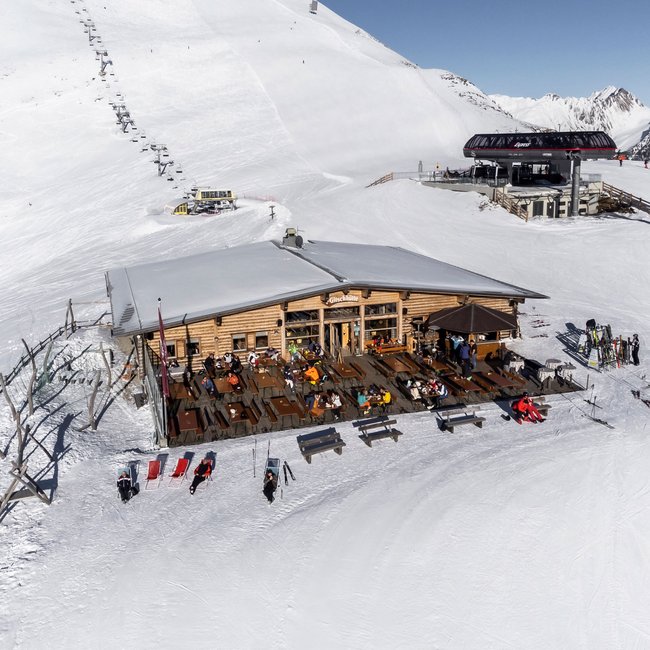 Gitschhütte Eine Berghütte mit Terrasse steht auf einem schneebedeckten Berg, umgeben von Skifahrern und Sesselliften. Menschen sitzen draußen und genießen das sonnige Wetter.