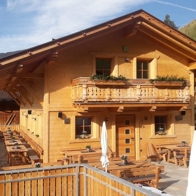 Hofschänke Lechnerhof Een berghut van licht hout met een balkon en bloembakken staat op een berghelling. Voor de hut staan meerdere houten tafels en banken met parasols op een terras.