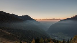 Pfunders: urig und idyllisch Das Bild zeigt eine weitläufige Berglandschaft während des Sonnenaufgangs oder Sonnenuntergangs. Nebel füllt die Täler zwischen den Bergen, während die Gipfel von weichem, goldenem Licht beleuchtet werden, was eine friedliche und malerische Atmosphäre erzeugt.