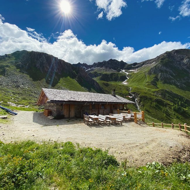Kröllhütte-Eisbruggalm A mountain cabin made of wood and stone stands on a sunny, green mountain meadow, surrounded by tall, rocky mountains. In front of the cabin, there are several wooden tables and benches.