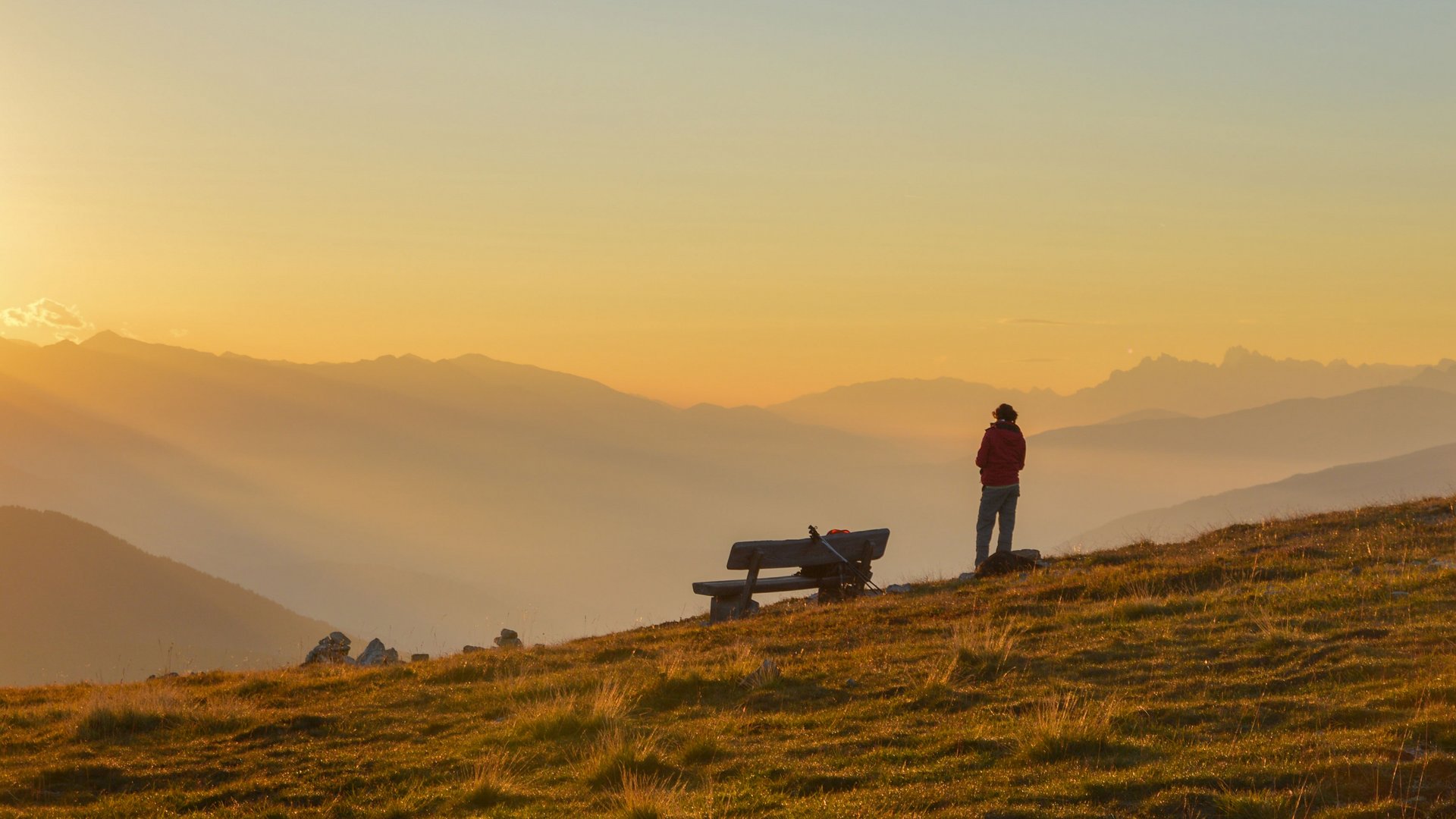 Vals - Jochtal - Steinermandl The picture shows a person standing on a grassy mountain meadow, looking into the distance as the sun sets on the horizon, casting the sky in warm yellow tones. Next to the person is a wooden bench, and in the background, gentle, mist-covered mountain ranges can be seen.