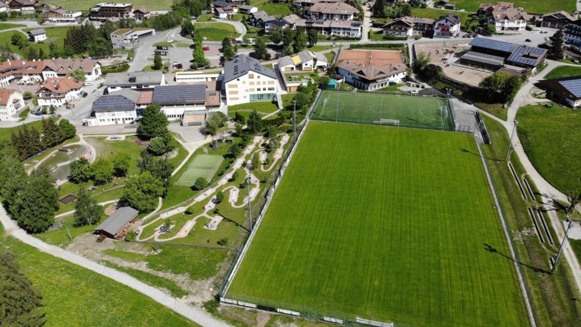 Het Aktivpark Terenten De afbeelding toont een luchtfoto van een dorp met een grote groene weide die dient als voetbalveld. Rond het veld bevinden zich verschillende gebouwen, waaronder huizen met zonnepanelen op de daken, een midgetgolfbaan en paden die door een verzorgd, groen landschap lopen.