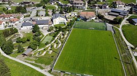Het Aktivpark Terenten De afbeelding toont een luchtfoto van een dorp met een grote groene weide die dient als voetbalveld. Rond het veld bevinden zich verschillende gebouwen, waaronder huizen met zonnepanelen op de daken, een midgetgolfbaan en paden die door een verzorgd, groen landschap lopen.