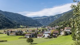 Vintl im Pustertal Das Bild zeigt eine malerische Landschaft mit grünen Wiesen und einem Dorf im Tal. Im Hintergrund sind bewaldete Hügel zu sehen, und in der Mitte des Dorfes ragt eine Kirche mit einem hohen, spitzen Turm hervor.