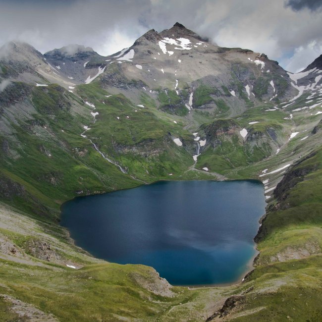 Wilde Kreuzspitze Das Bild zeigt einen hochgelegenen Bergsee, umgeben von steilen, grünen Berghängen und schneebedeckten Gipfeln. Der Himmel ist wolkenverhangen, was der Szenerie eine dramatische Atmosphäre verleiht.