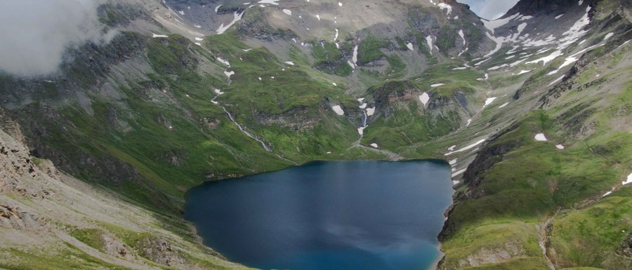 The Gitschberg webcams The picture shows a high-altitude mountain lake, surrounded by steep, green mountain slopes and snow-covered peaks. The sky is overcast, giving the scene a dramatic atmosphere.