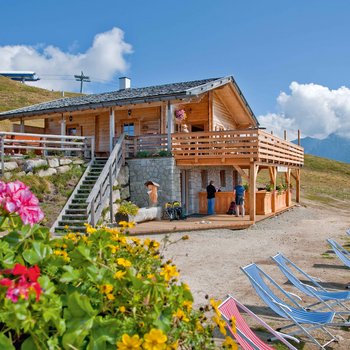 Südtiroler Almkost Das Bild zeigt eine gemütliche Berghütte aus Holz mit einer großen Terrasse und Blumenkästen. Im Vordergrund sind bunte Blumen und mehrere Liegestühle zu sehen. Menschen stehen und unterhalten sich vor der Hütte. Im Hintergrund sind grüne Hügel und Berge unter einem blauen Himmel mit wenigen Wolken.