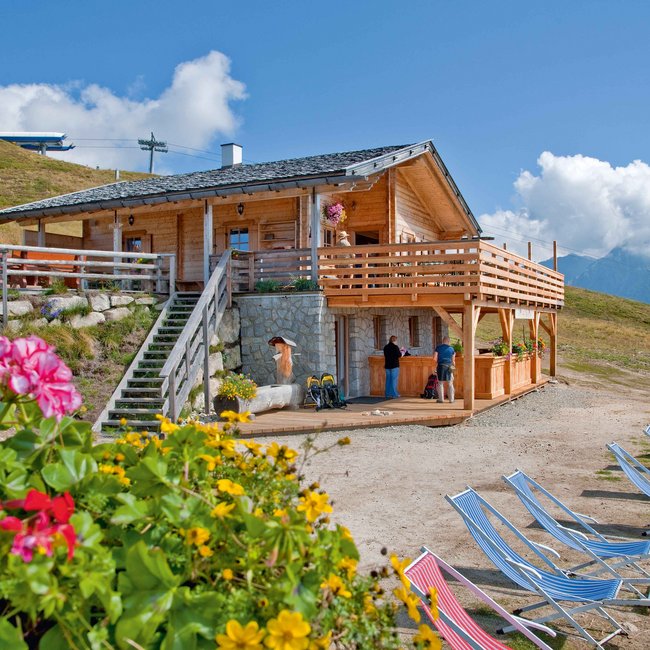 Ochsenalm Das Bild zeigt eine gemütliche Berghütte aus Holz mit einer großen Terrasse und Blumenkästen. Im Vordergrund sind bunte Blumen und mehrere Liegestühle zu sehen. Menschen stehen und unterhalten sich vor der Hütte. Im Hintergrund sind grüne Hügel und Berge unter einem blauen Himmel mit wenigen Wolken.